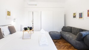 A modern hotel room with a bed, a gray sofa, a wooden coffee table, and a white wall with framed artwork.