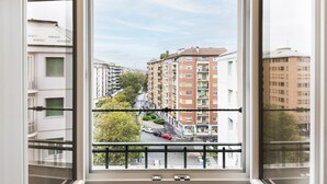 A view from a balcony with a metal railing, overlooking a street with multi-story buildings, cars, and trees.