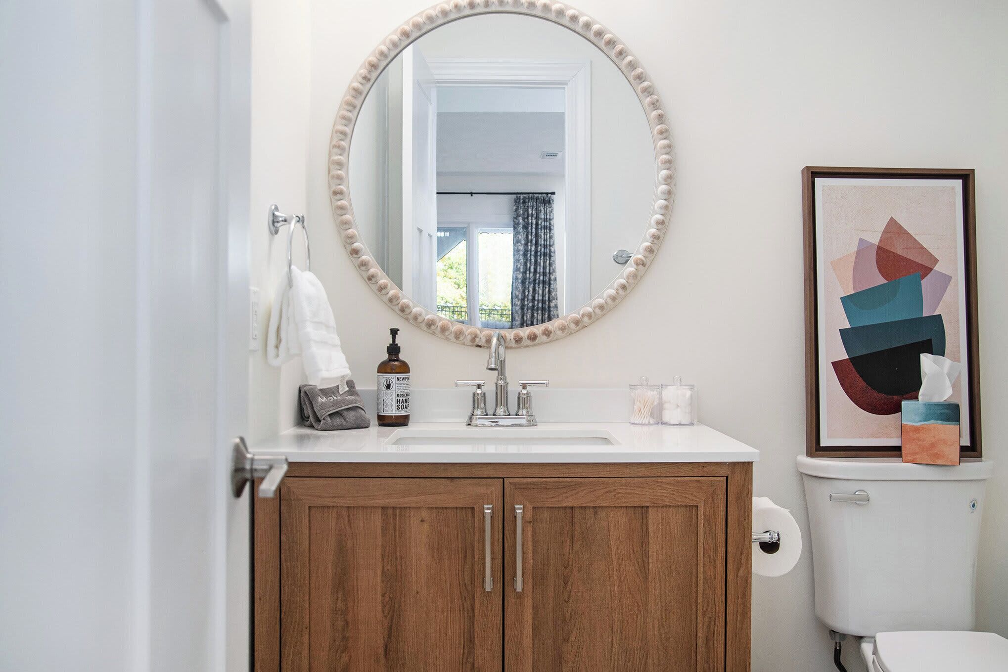 A bathroom with a wooden vanity, a round mirror with a beaded frame, a white sink, and a toilet.