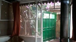 Una habitación con una silla, una mesita y una puerta corrediza entreabierta. Se observa un portón verde en el exterior.