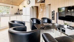 A modern kitchen with white cabinets, a black countertop, and a built-in stove. There is a dining area with black leather chairs and a glass table. A large TV is mounted on the wall, and a clock is visible on the wall. - 5 Star Holiday Home in Idestrup (Idestrup)