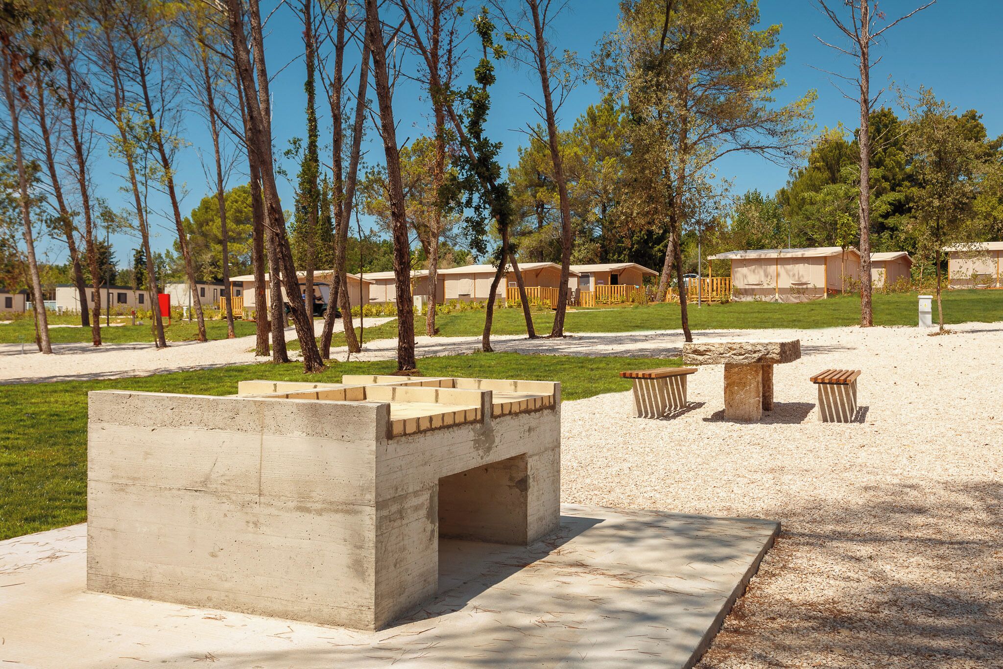 A concrete structure with benches and a stone table in a park-like setting with trees and a row of mobile homes in the background.
