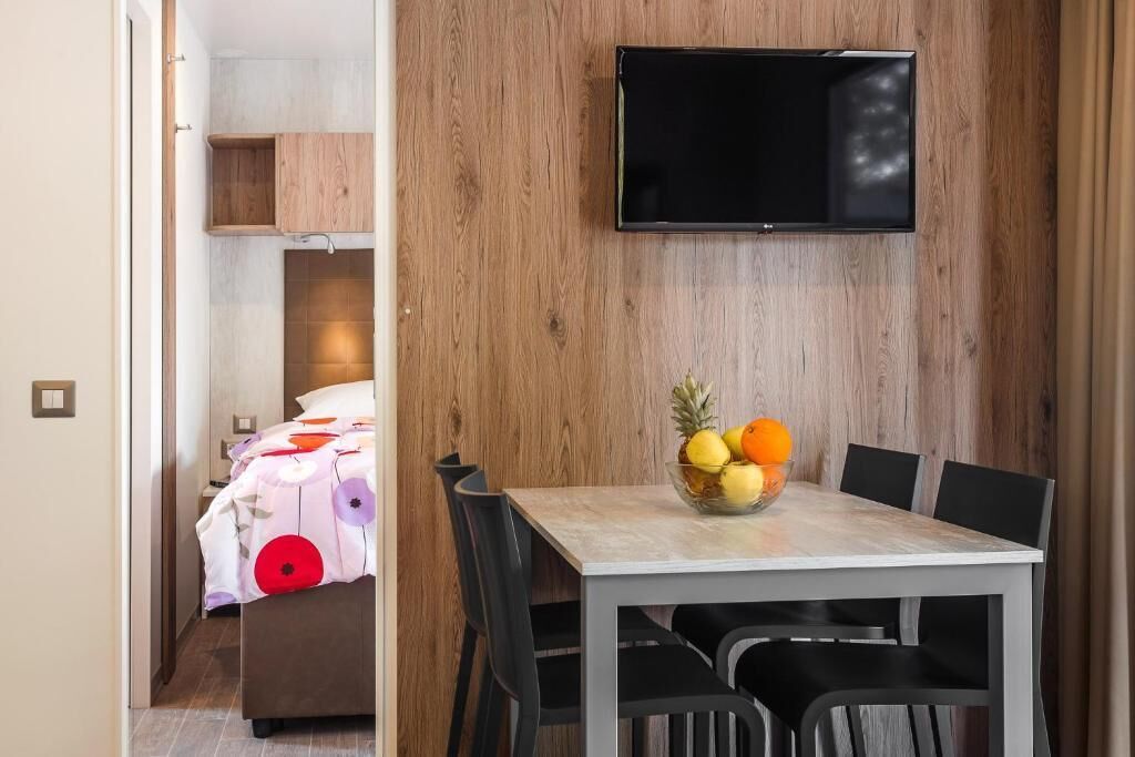 Ein Hotelzimmer mit einer Holzwand, einem flachbildfernseher über einem Esstisch mit Obstschale und einem Bett mit blumigem Überwurf.