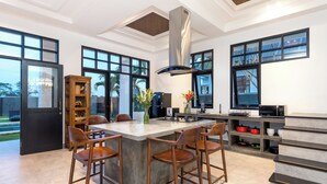 A modern kitchen with a central island, bar stools, a built-in bookshelf, and large windows. - Villa Sakayana (Canggu)