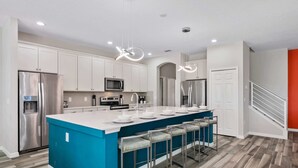 Una cocina moderna con armarios blancos, electrodomésticos de acero inoxidable, una isla azul con taburetes de bar y una escalera con una pared de acento roja.