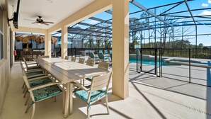 Un amplio espacio exterior para comer con una larga mesa y sillas, un ventilador de techo y vistas a una piscina.