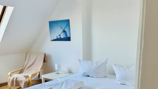 Ein Hotelzimmer mit einem Doppelbett, einem Sessel, einem kleinen Tisch und einem Windmühlenbild an der Wand.