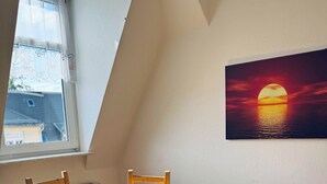 Ein Essbereich mit einem runden Tisch, bedeckt mit einer gelben Tischdecke, umgeben von Holzsesseln, einem Fenster mit Blick auf Gebäude und einem Sonnenuntergangsgemälde an der Wand.