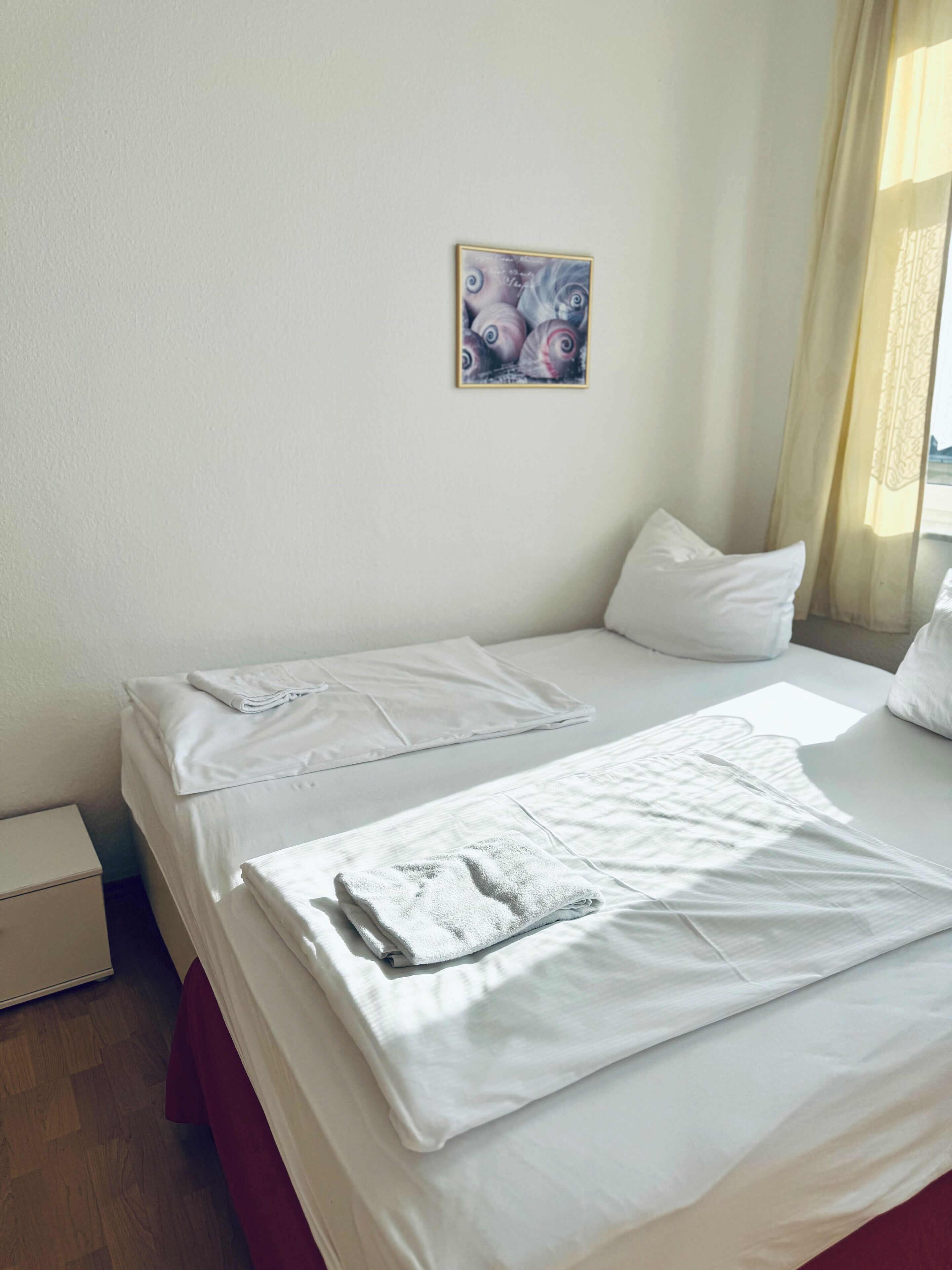 A neatly made bed with white linens and a folded towel, a small bedside table, and a painting on the wall.