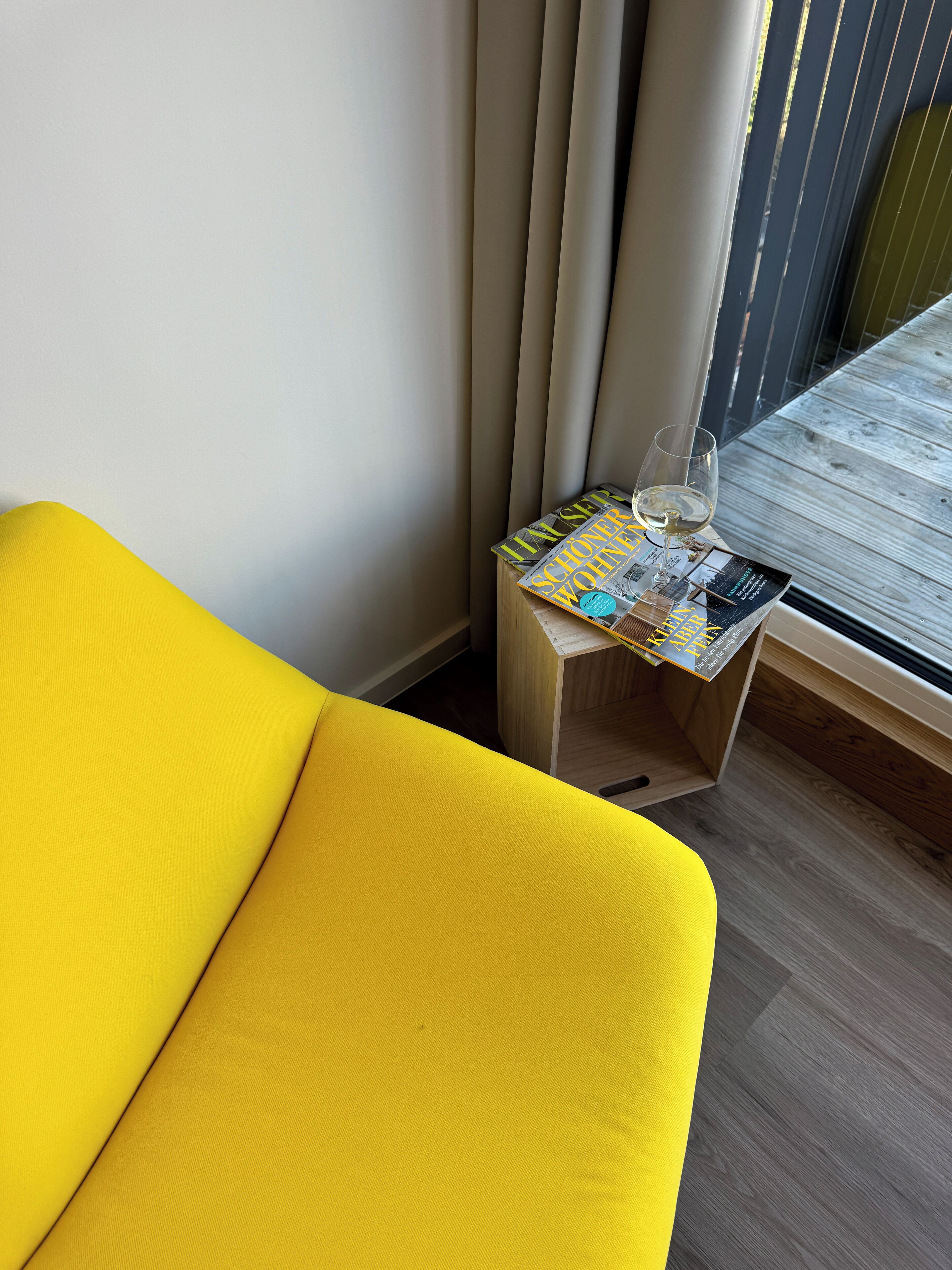 Ein gelbes Sofa, ein kleiner Holztisch mit einer Zeitschrift und einem Glas Weißwein, sowie ein Fenster mit vertikalen Jalousien.