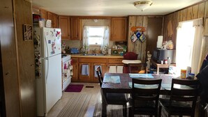 A compact kitchen with wooden cabinets, a refrigerator, a dining table with chairs, and a window with curtains.