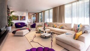 A modern living room with a beige sofa, a purple armchair, and a coffee table. There are large windows with curtains, a plant, and artwork on the walls. - Villa Blanchette  (Marrakech)
