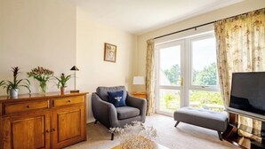 Ein Wohnzimmer mit einer Holzkommode, einem grauen Sessel, einem kleinen Hocker, einem Couchtisch mit getrockneten Blumen, einem Fernseher und einem großen Fenster mit Vorhängen.