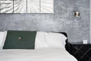 A bed with white linens and a green pillow, a black nightstand with a white object on it, and a wall with a textured pattern and a wall-mounted light fixture.
