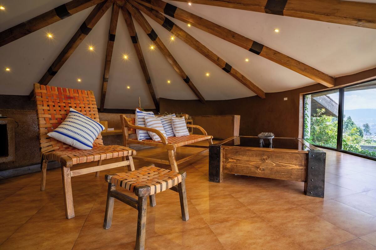 A high-ceilinged room with wooden beams, a large window, and a seating area with wicker chairs and cushions.