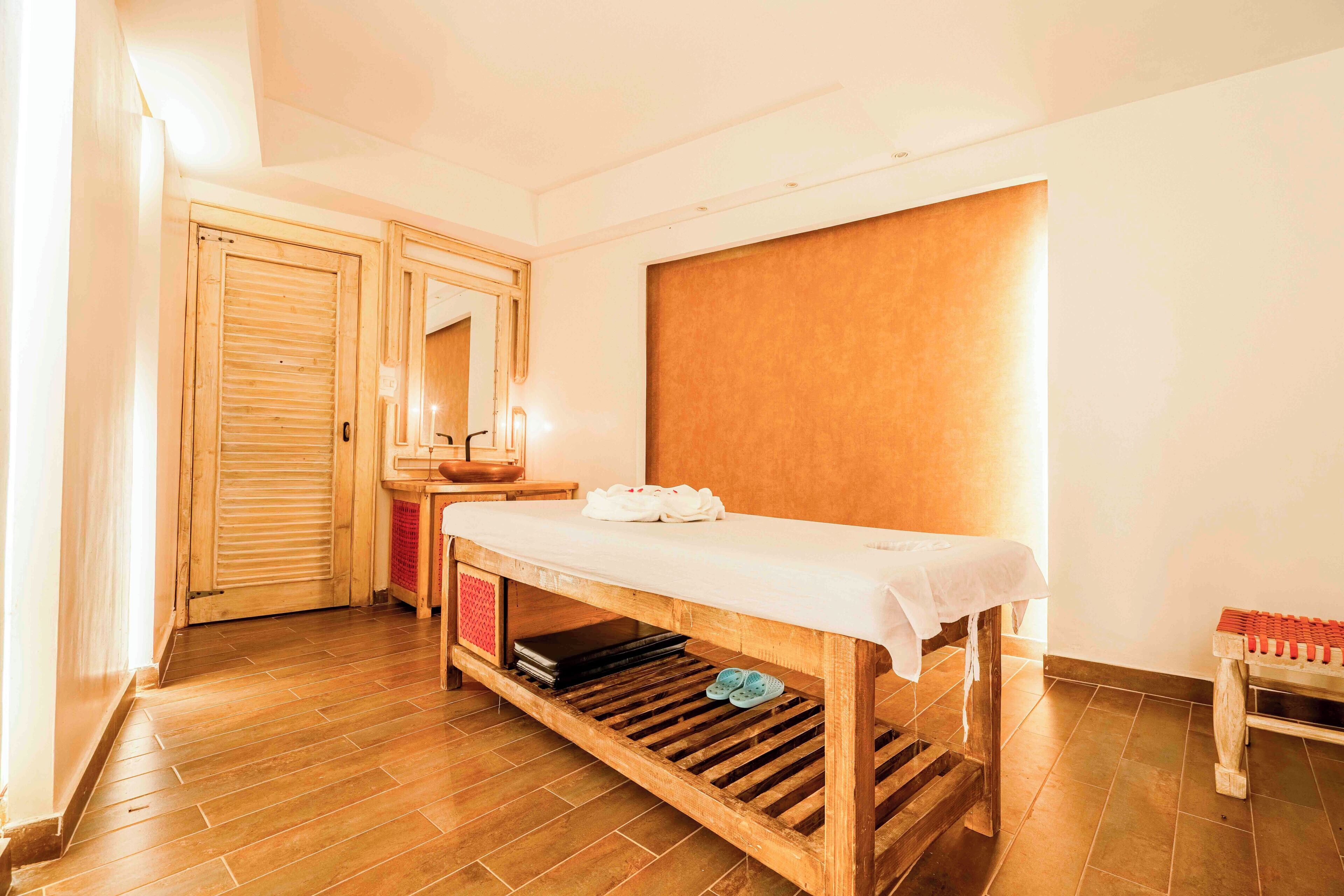 A wooden massage table with a white sheet, a wooden bench, and a small shelf with a red object.