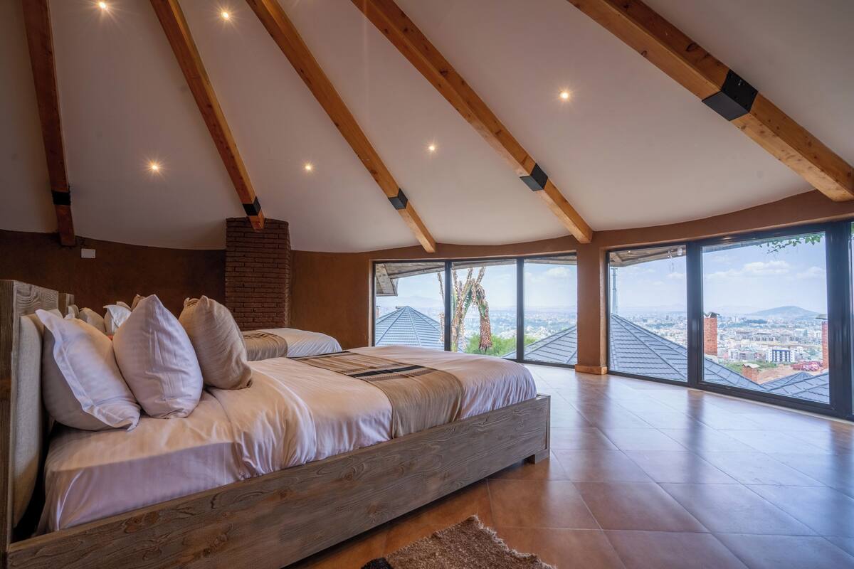 A bedroom with a large bed, wooden beams, a brick fireplace, and a city view through large windows.