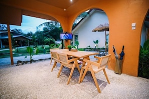 An outdoor dining area with a wooden table and chairs, a television on a wall, and a view of a house with a thatched roof. - Lilla Rainforest Retreat @ Hulu Langat (Hulu Langat)