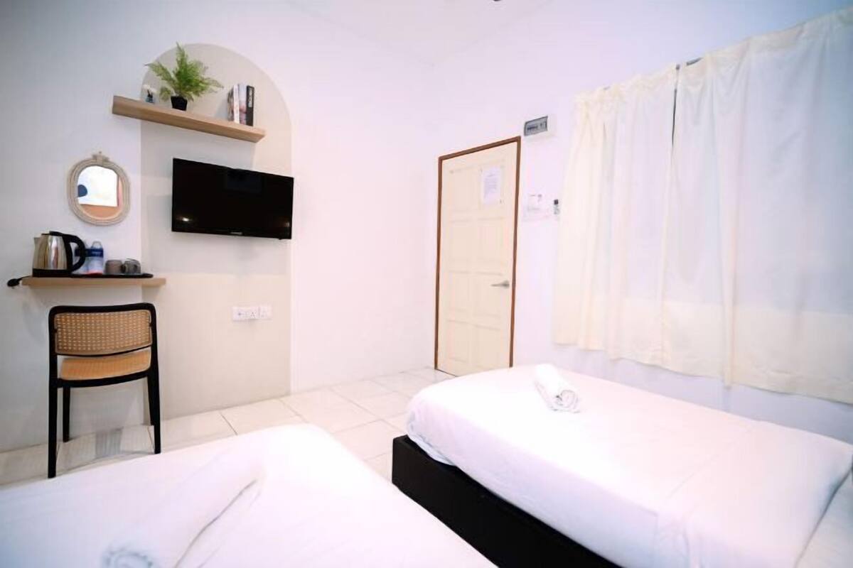 A hotel room with two beds, a TV, a chair, a shelf with a plant, and a door.