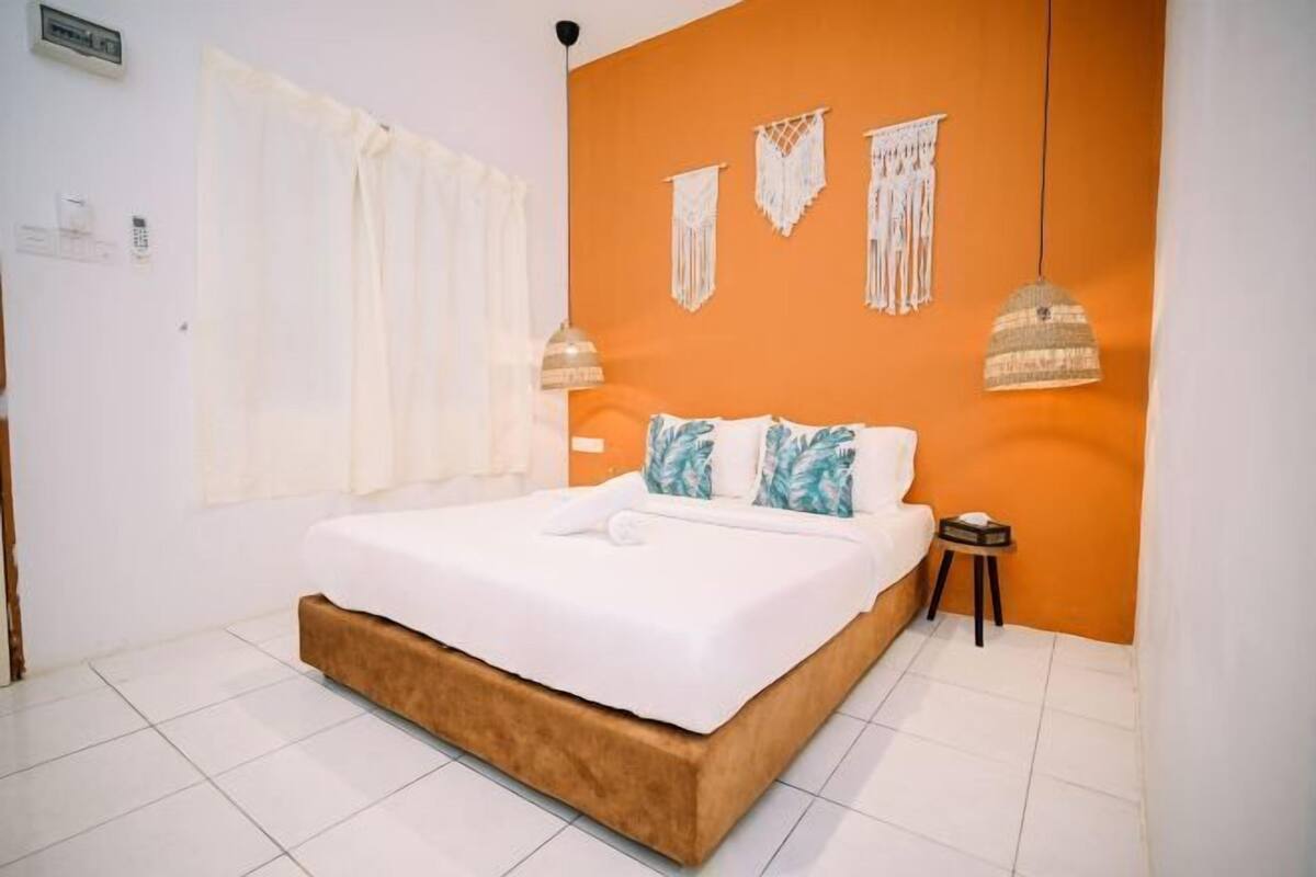A bedroom with a bed, white curtains, an orange wall, and a small bedside table.