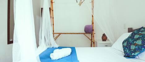 A room with a bed, a mosquito net, a wooden shelf, and a small table with a vase.