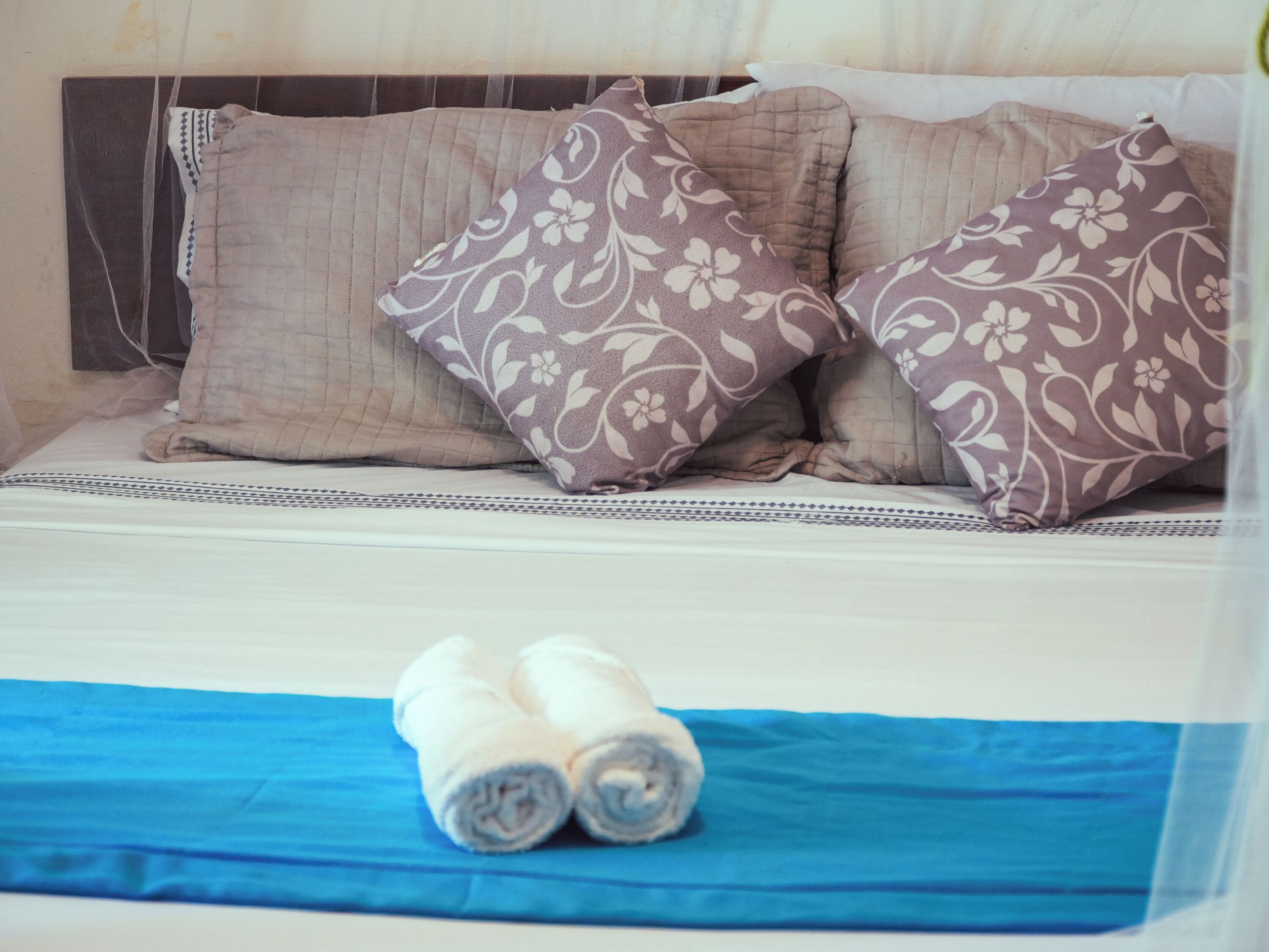 A bed with two pillows, a blue blanket, and two rolled towels.