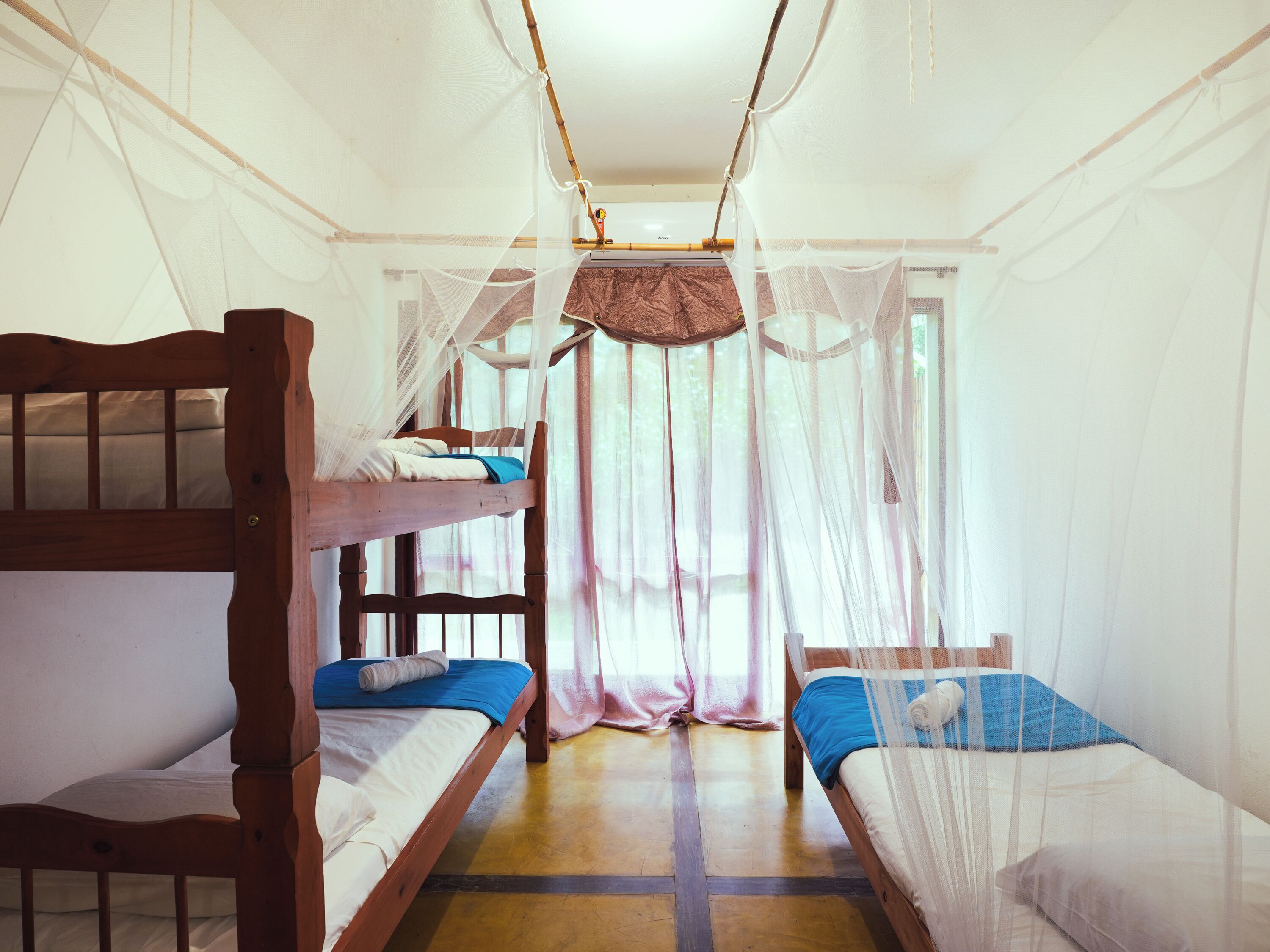 A room with two wooden bunk beds, a mosquito net, and a window with sheer curtains.