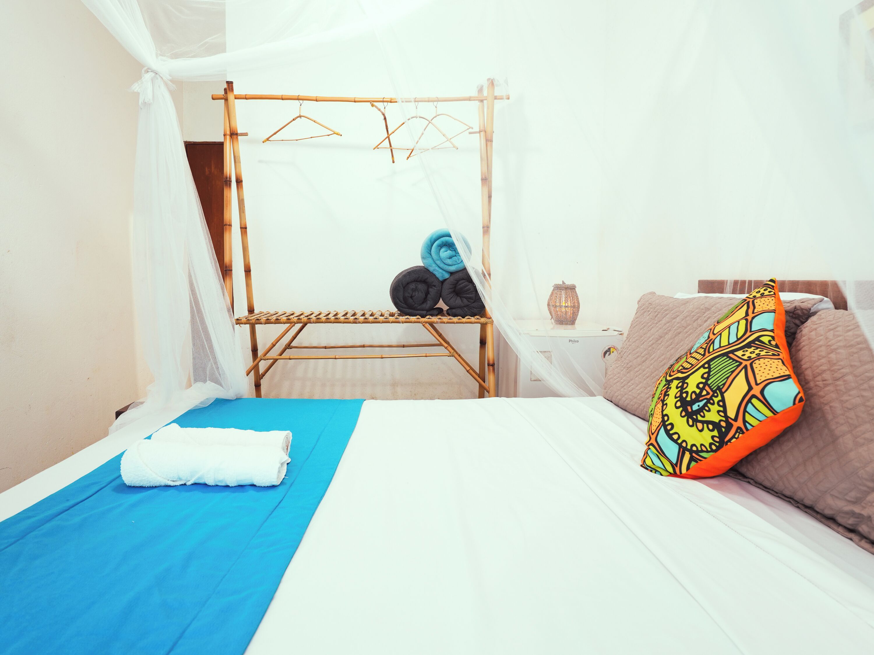 Une chambre à coucher avec un lit, un étendoir à linge suspendu, une table de chevet avec une lampe et un coussin coloré.