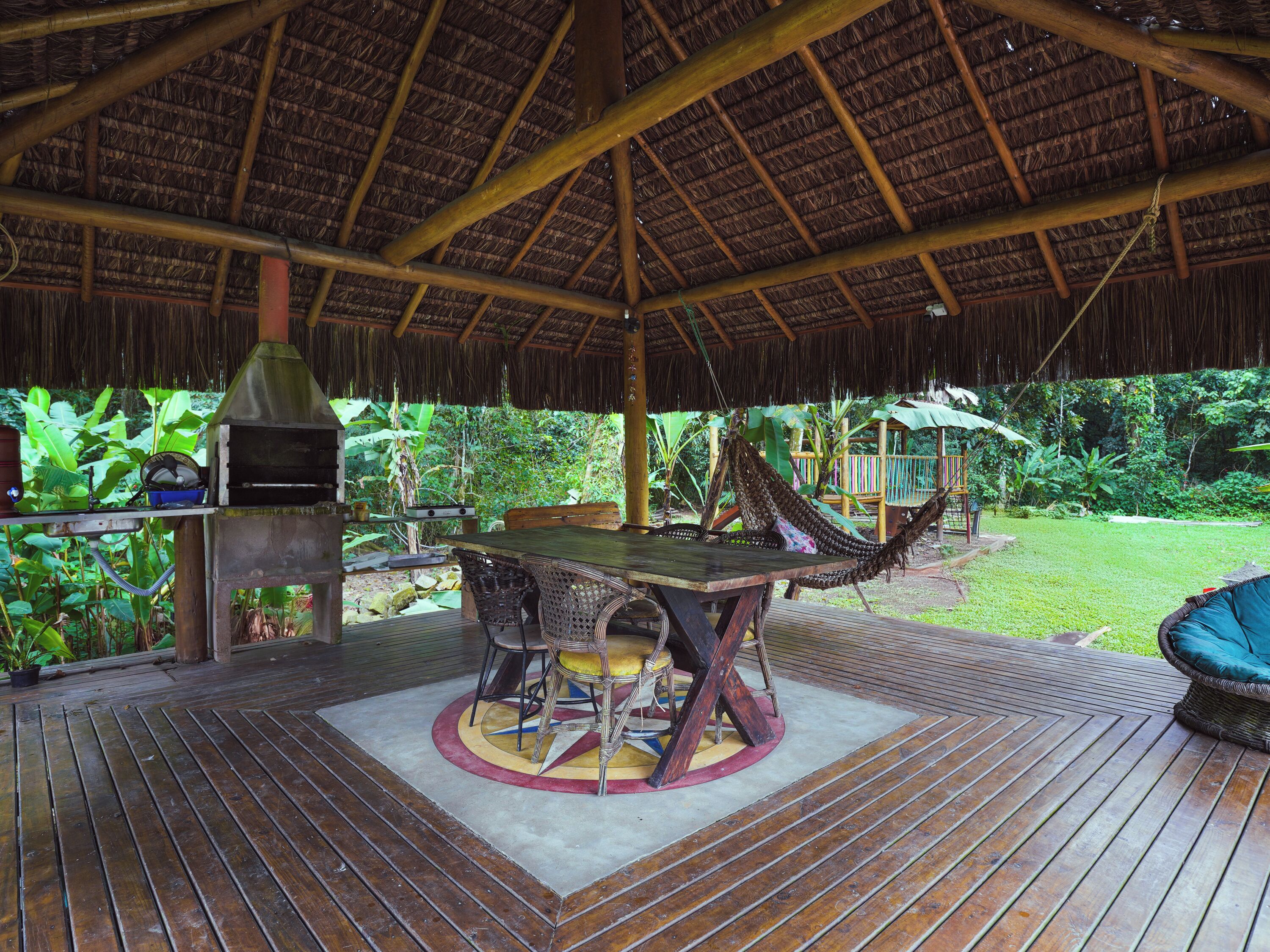 Un espacio cubierto para comer al aire libre, con una mesa y sillas de madera, una estación de parrillada y una hamaca.
