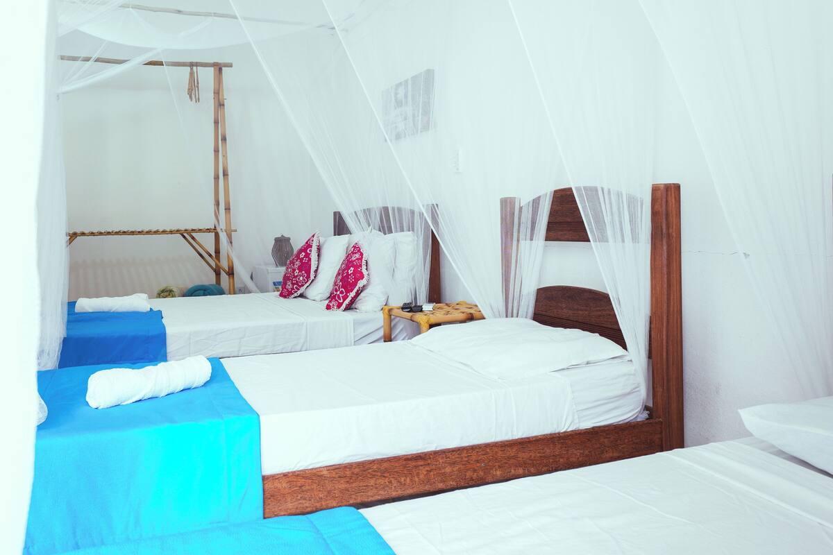 Ein Zimmer mit zwei Betten, jeweils mit weißer Bettwäsche und einem Holz-Kopfteil. Über den Betten ist ein Moskitonetz gespannt, und dazwischen steht ein kleiner Tisch mit einer Lampe.