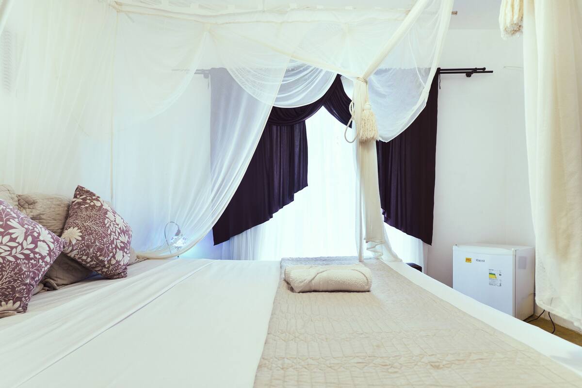 Ein ordentlich bezogenes Bett mit weißer Decke und beiger Steppdecke, ein Baldachin mit weißen Vorhängen und ein kleiner Nachttisch mit weißer Lampe.