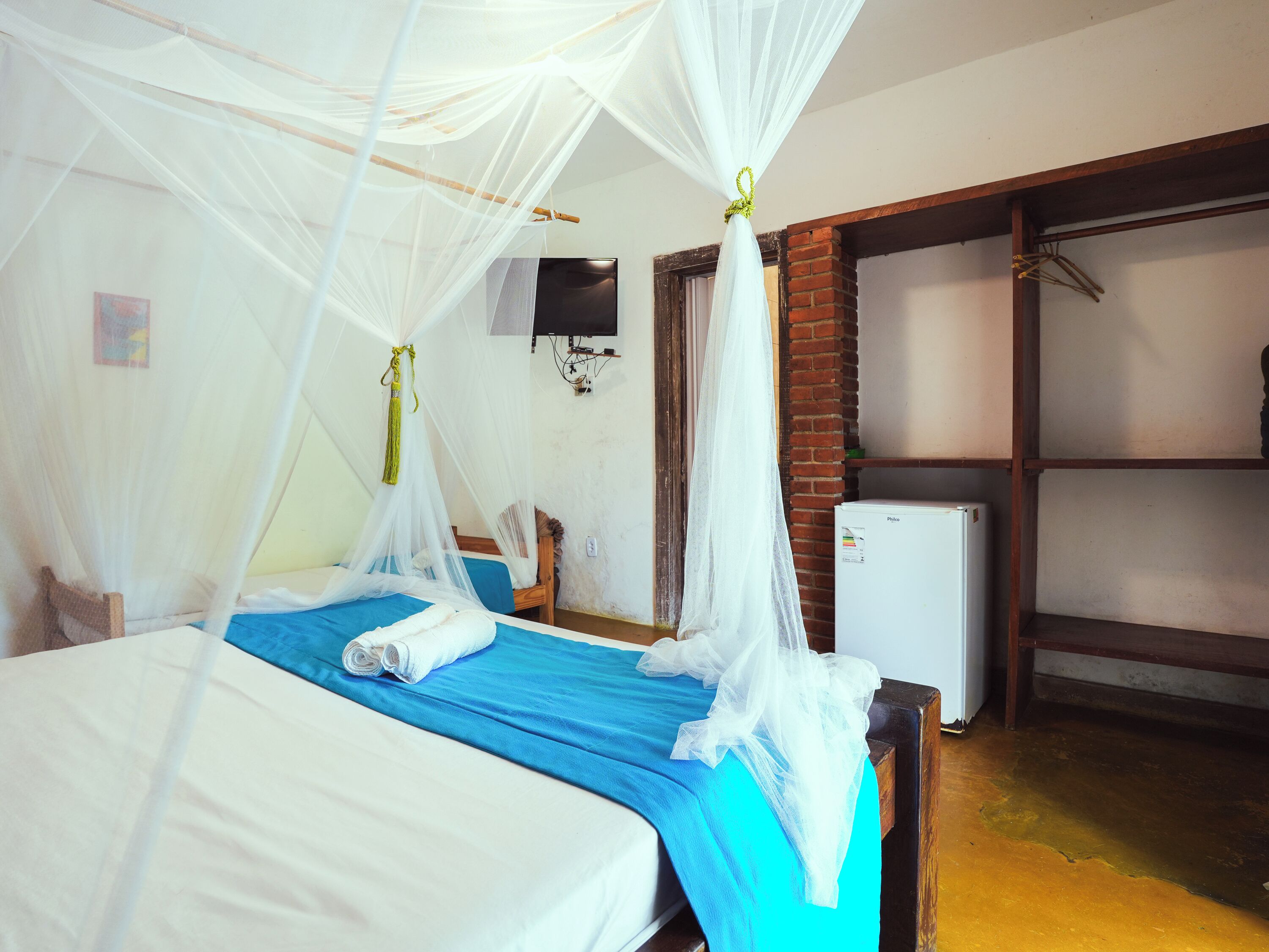 Una habitación con una cama cubierta por una mosquitera, un edredón azul y un colchón blanco. Hay un refrigerador pequeño y un televisor montado en la pared.