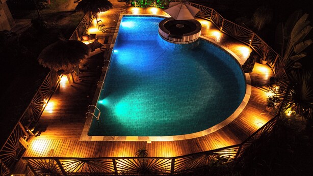 A nighttime view of a swimming pool with a wooden deck, lounge chairs, and a thatched-roof structure. - VELINN Pousada Bali Itamambuca (Ubatuba)
