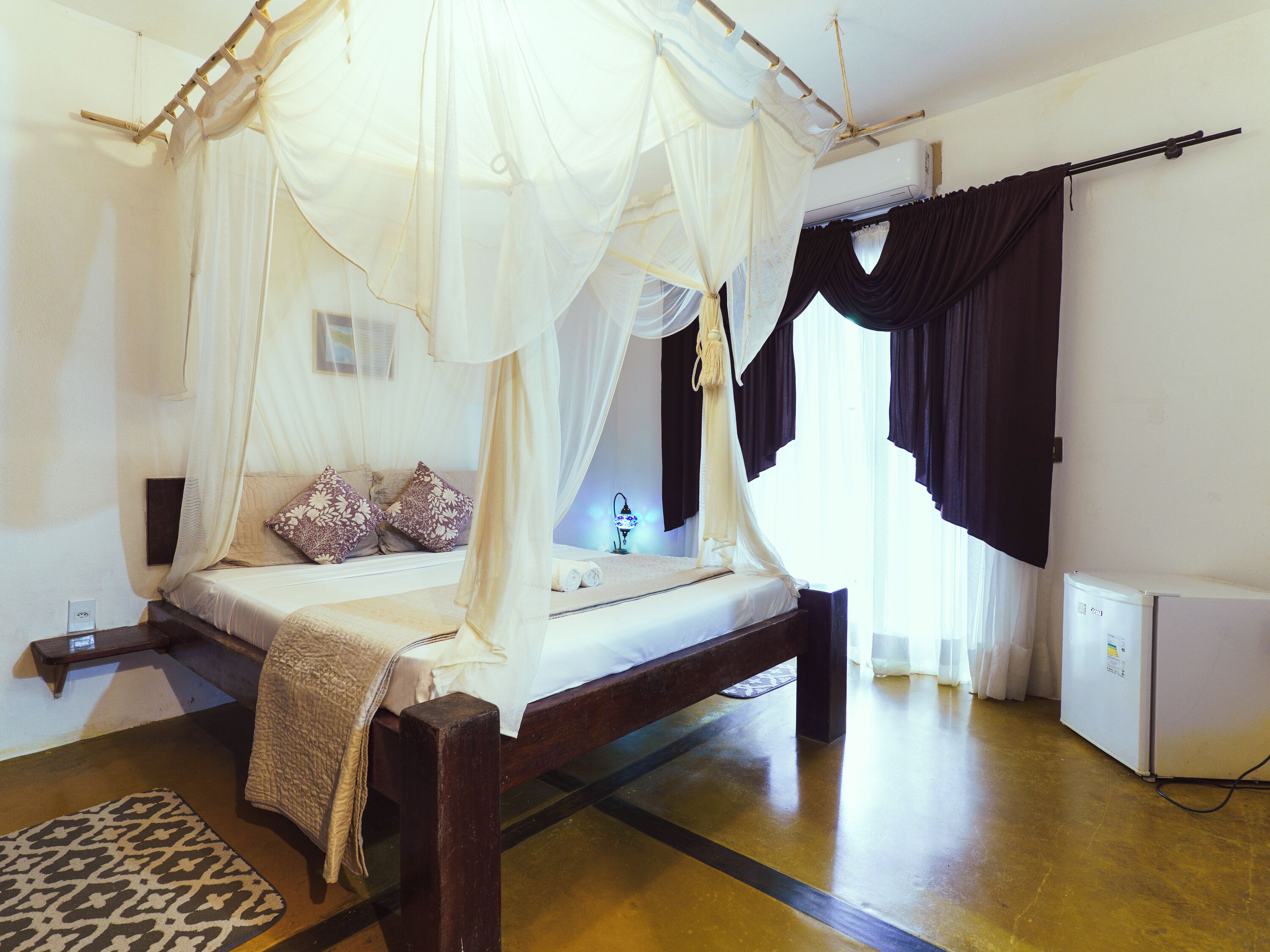 A room with a bed under a canopy, a small table with a lamp, and a refrigerator.