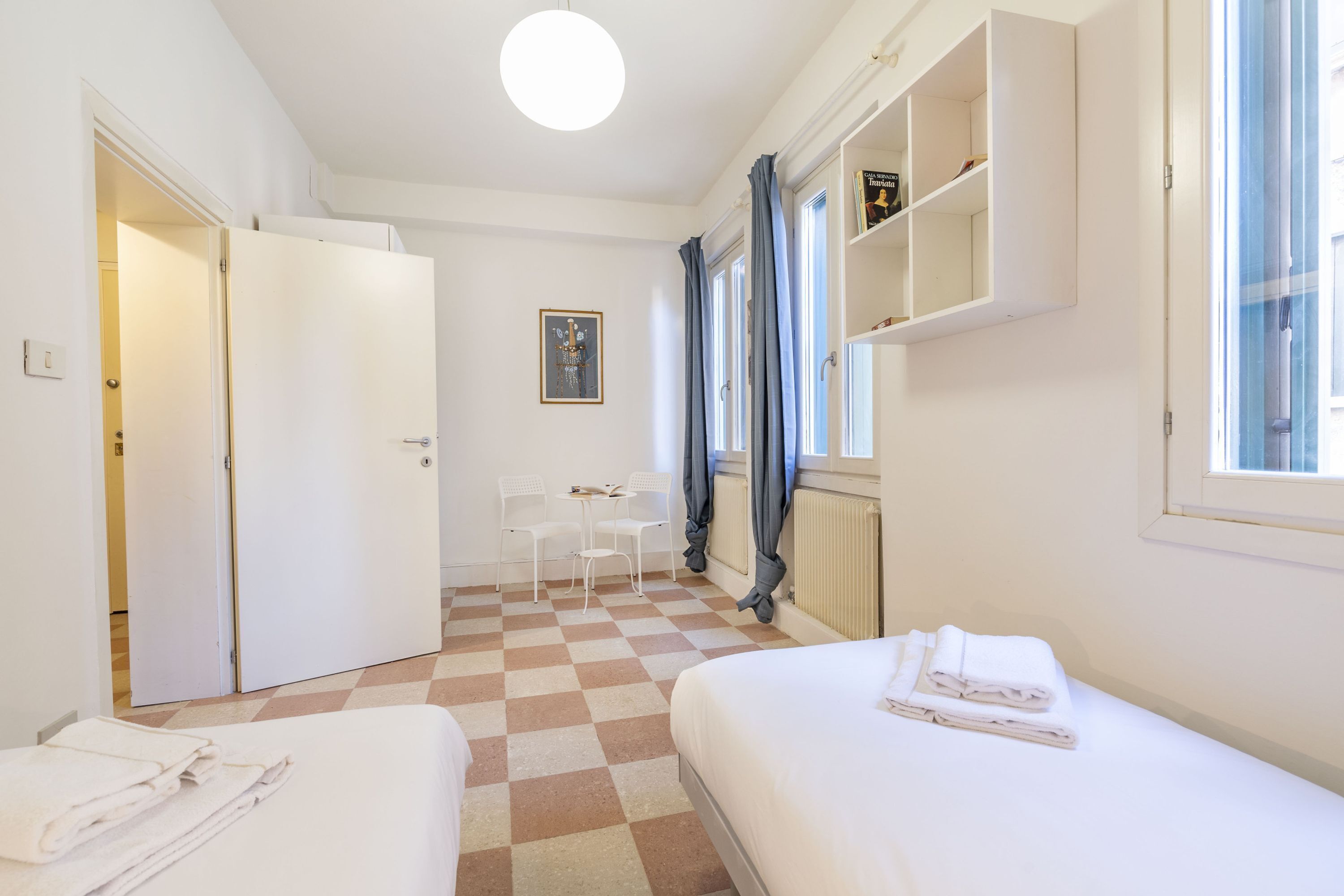 Una camera d'albergo con due letti, un pavimento a scacchi, una scrivania con sedie e una finestra con tende.