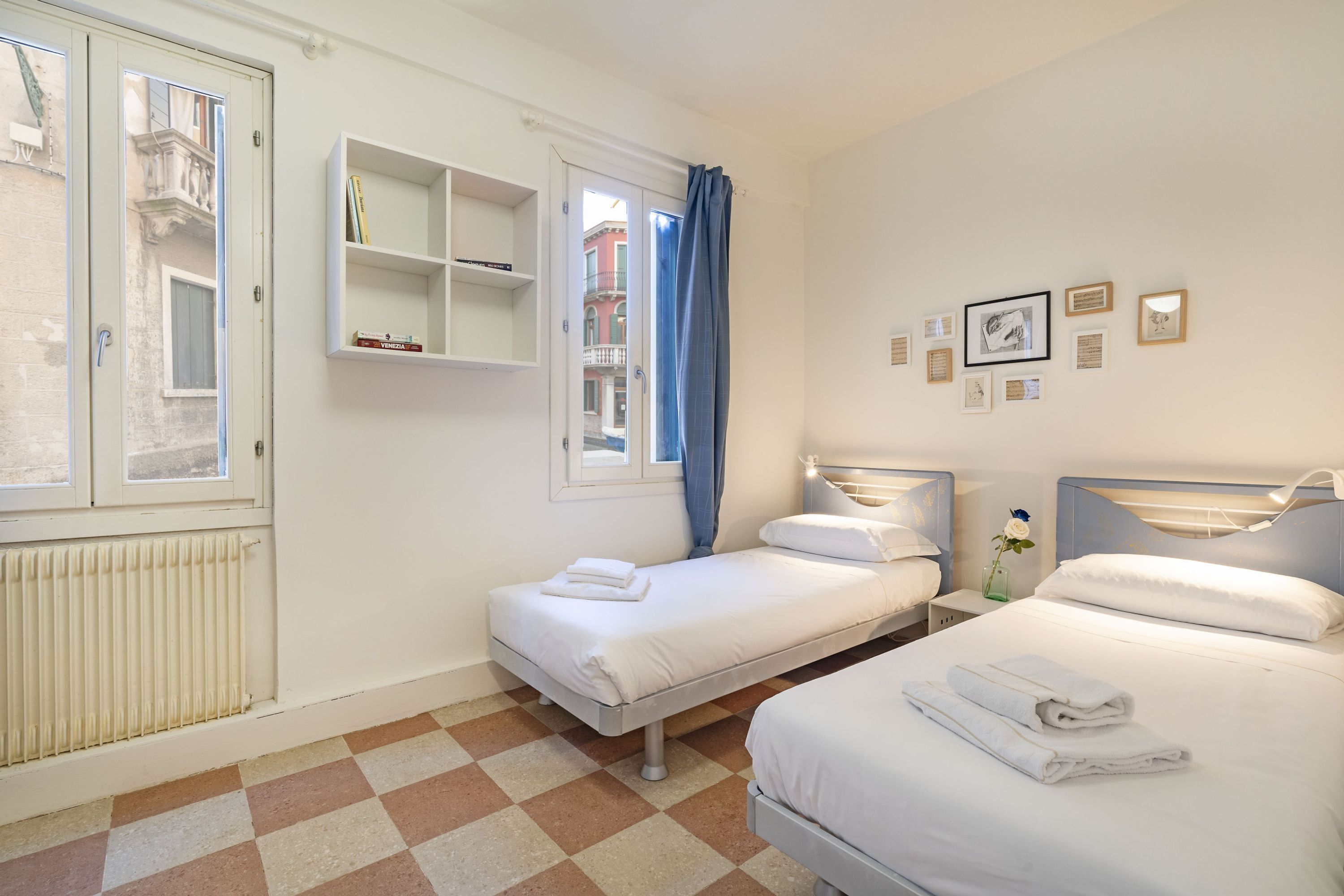 A hotel room with two beds, a radiator, a checkered floor, a window with a curtain, and a wall with framed pictures.