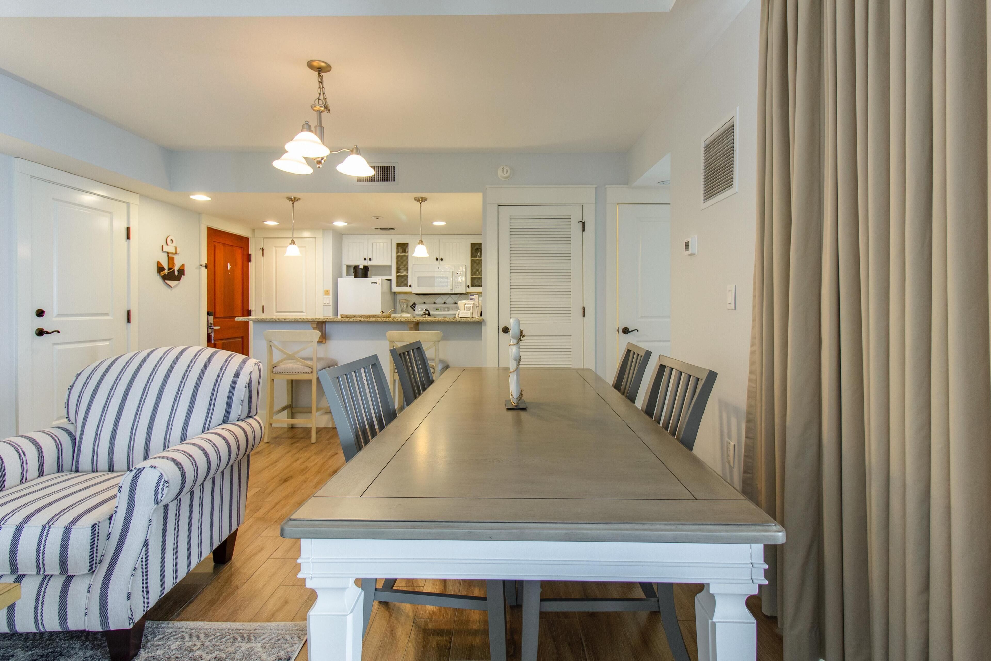 A dining area with a long table, striped armchairs, and a kitchen in the background.