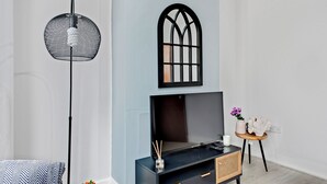 Una sala de estar moderna con televisor, un adorno en la pared, una lámpara de pie y una pequeña mesa con un florero y una vela.