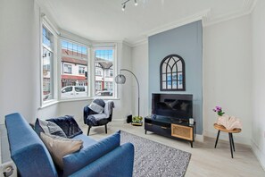 A modern living room with a large window, a flat-screen TV, a sofa, and a small table with a vase. - Lovely Home - Park Views & Ideal Transport Links (London)
