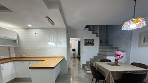 Una cocina moderna con encimera de madera y armarios blancos, un comedor con mesa y sillas, y una escalera que sube a otro nivel.