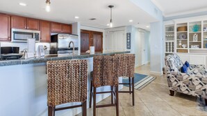 A modern kitchen with stainless steel appliances, a bar area with woven chairs, and a patterned sofa in a bright living room.
