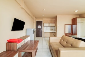 A modern living room with a wooden table, a beige sofa, a flat-screen TV mounted on the wall, and a kitchen area in the background.