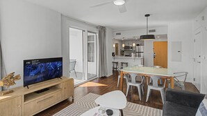 A modern living room with a flat-screen TV, a dining area with a glass table, and a kitchen in the background. - Elation 2BR: Sunset Views & Steps to Baytowne (Miramar Beach)