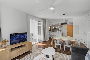 Una sala moderna con televisor de pantalla plana, un comedor con mesa de vidrio y una cocina al fondo.