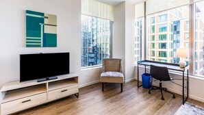 A modern living room with a flat-screen TV, a comfortable chair, a desk with a lamp, and large windows offering a city view. - Seaport Apt with City Views and Pool (Boston)