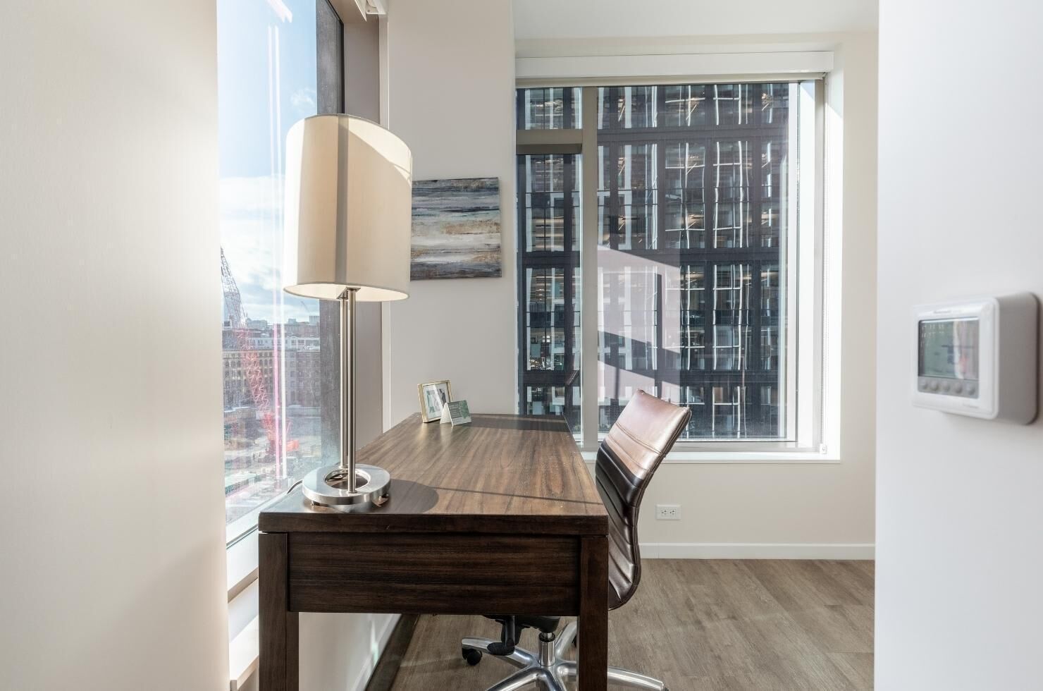 Un espacio de oficina moderno con un escritorio de madera, una lámpara, una silla y vistas de un paisaje urbano a través de amplios ventanales.