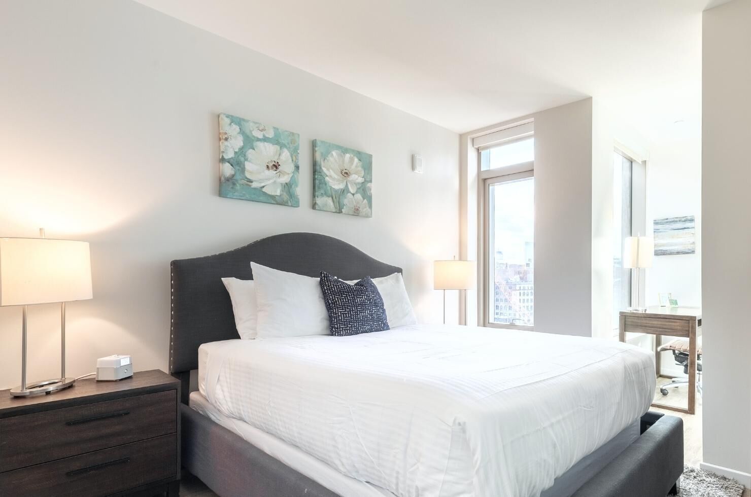 Une chambre d’hôtel avec un grand lit, deux lampes de chevet, une table de nuit, une fenêtre donnant sur la ville et deux œuvres d’art florales accrochées au mur.