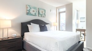 Hotellihuone, jossa on suuri sänky, kaksi yöpöytälamppua, yöpöytä, ikkuna, josta on näkymä kaupunkiin, sekä kaksi kukka-aiheista taideteosta seinällä.