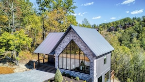 A modern cabin with a sloped roof, large windows, and a stone facade, surrounded by a forested landscape. - WALDEN'S RIDGE SPLASH! LUXURY 2 BEDROOMS! (Sevierville)