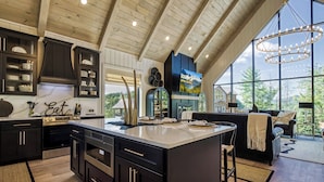 A modern kitchen with dark cabinetry, a large island, and a view of the outdoors through large windows. - WALDEN'S RIDGE SPLASH! LUXURY 2 BEDROOMS! (Sevierville)